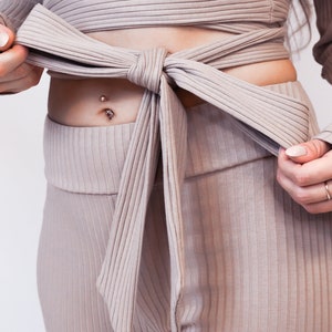 May include: A ribbed knit top and pants set in a light brown color. The top is a wrap style with a long sleeve and a tie at the waist. The pants are high-waisted and have a straight leg.