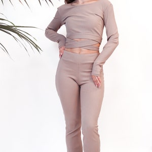 May include: A beige ribbed long-sleeve top with a wrap detail at the waist and matching ribbed pants. The outfit is styled with white sneakers.