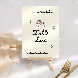 May include: A white card with black calligraphy that reads "Table Six" and a pink cake illustration. The card is on a small wooden easel.