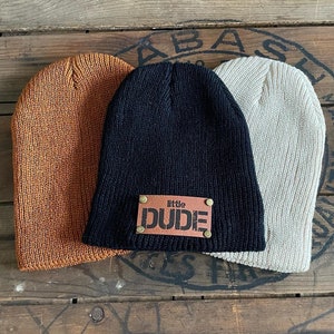 May include: Three knitted beanies in different colors. One beanie is black with a brown leather patch that says "little DUDE".