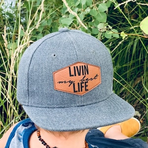 Puede incluir: Gorra de béisbol gris con visera plana y parche de cuero marrón con la inscripción "LIVIN my best LIFE". La gorra la lleva un niño. El niño lleva una camiseta sin mangas azul y gris con estampado de dinosaurios.