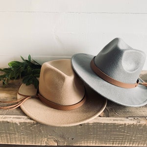 May include: Two felt fedora hats with leather bands and tassels. The hat on the left is tan and the hat on the right is grey.