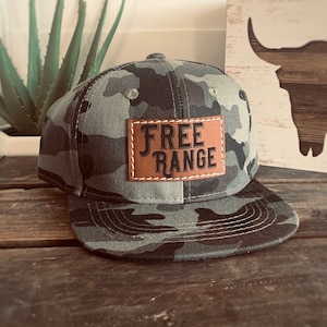 May include: Camouflage print baseball cap with a flat brim. A brown leather patch on the front displays the words "FREE RANGE" in a bold font. The cap is set on a rustic wooden surface.