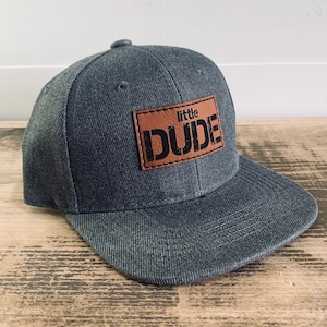 Pode incluir: Um boné de beisebol cinza escuro com um remendo de couro marrom que diz "little DUDE" em letras pretas.