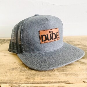 May include: Grey and black mesh trucker hat with a brown leather patch that says "big DUDE" in black lettering.