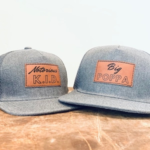 May include: Two gray trucker hats with brown leather patches. The smaller hat has the text "Notorious K.I.D." on the patch. The larger hat has the text "Big Poppa" on the patch.