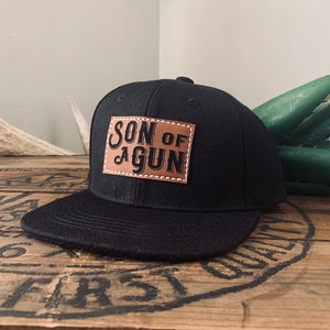 Op de afbeelding: Zwarte baseballpet met een bruin leren patch waarop in zwarte letters "Son of a Gun" staat.