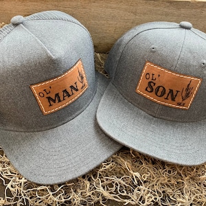 Op de afbeelding: Twee grijze baseballpetten met bruine leren patches. De patch op de linker pet zegt "Ol' Man" en de patch op de rechter pet zegt "Ol' Son".