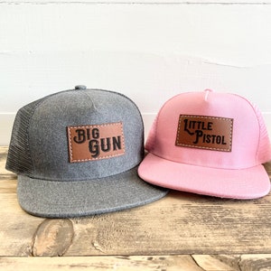 以下が含まれることがあります： 革のパッチが付いたトラッカーハット2つ。グレーの帽子には、黒い文字で「BIG GUN」と書かれた茶色の革のパッチが付いています。ピンクの帽子には、黒い文字で「LITTLE PISTOL」と書かれた茶色の革のパッチが付いています。
