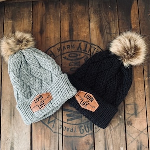 Puede incluir: Dos gorros de invierno de punto con pompones de piel sintética. Los gorros son grises y negros con un parche de cuero marrón que dice "Livin' my best LIFE".