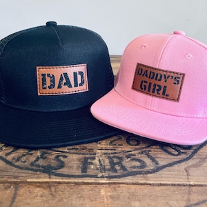 Könnte beinhalten: Zwei Kappen: eine schwarze Trucker-Kappe mit einem braunen Lederaufnäher mit der Aufschrift "DAD" und eine rosa Baseballkappe mit einem braunen Lederaufnäher mit der Aufschrift "DADDY'S GIRL".