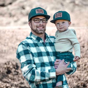 May include: Two dark green baseball caps featuring a rectangular patch with the text "BLAME ROOTS" in a stylised font. The caps have a structured crown and a flat brim, designed with a matching dark green fabric and a consistent logo patch.