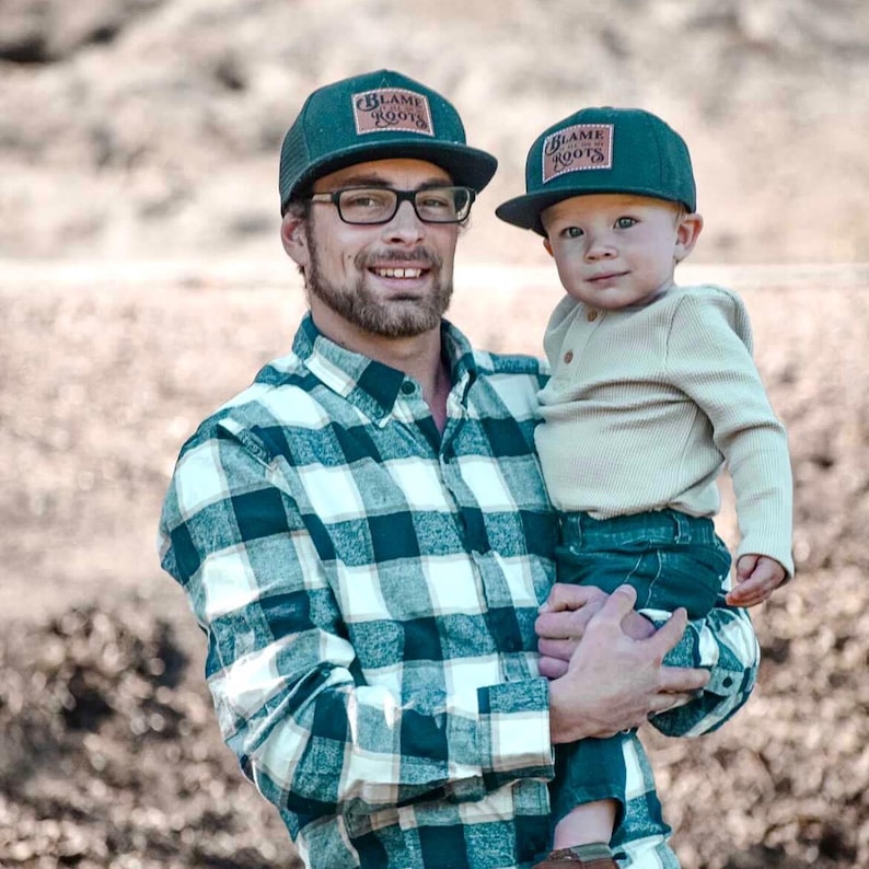 May include: Two dark green baseball caps featuring a rectangular patch with the text "BLAME ROOTS" in a stylised font. The caps have a structured crown and a flat brim, designed with a matching dark green fabric and a consistent logo patch.