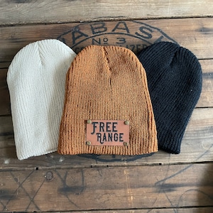 May include: Three knit beanies in different colors. One is beige, one is brown with a leather patch that says "FREE RANGE", and one is black.