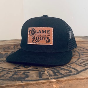 May include: A black trucker cap with a mesh back and a flat peak. Centred on the front is a rectangular tan faux leather patch featuring white stitched edges and the black text "BLAME IT ALL ON MY ROOTS" in a decorative, stylised font.