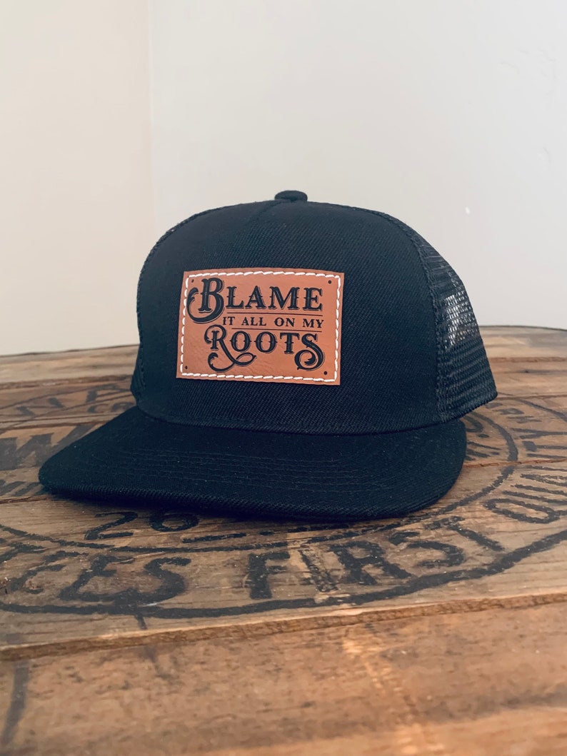 May include: A black trucker cap with a mesh back and a flat peak. Centred on the front is a rectangular tan faux leather patch featuring white stitched edges and the black text "BLAME IT ALL ON MY ROOTS" in a decorative, stylised font.