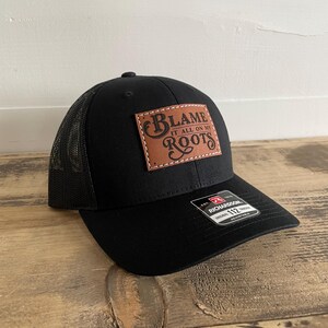 May include: A black trucker cap featuring a rectangular brown leather patch stitched onto the front panel. The patch displays the text "BLAME IT ALL ON MY ROOTS" in a decorative font. A black sticker on the brim reads "RICHARDSON 112".