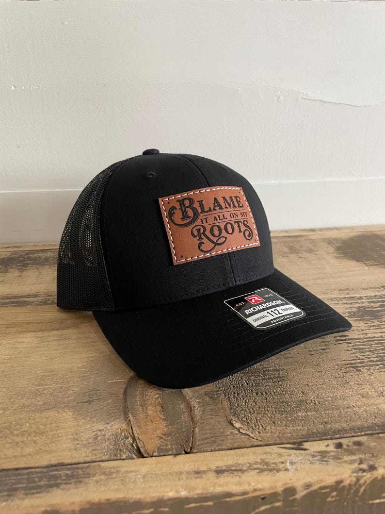 May include: A black trucker cap featuring a rectangular brown leather patch stitched onto the front panel. The patch displays the text "BLAME IT ALL ON MY ROOTS" in a decorative font. A black sticker on the brim reads "RICHARDSON 112".