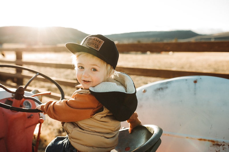 May include: A black baseball cap featuring a rectangular brown patch on the front with the text Blame Roots. The cap has a flat brim and is constructed from a solid black fabric, providing a structured look with clear, legible lettering.
