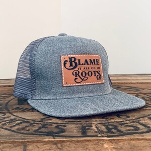 May include: A grey trucker hat with a mesh back and a flat brim. A rectangular tan leather patch is stitched to the front, featuring the black text "BLAME IT ALL ON MY ROOTS" in a decorative font with white stitching around the patch border.