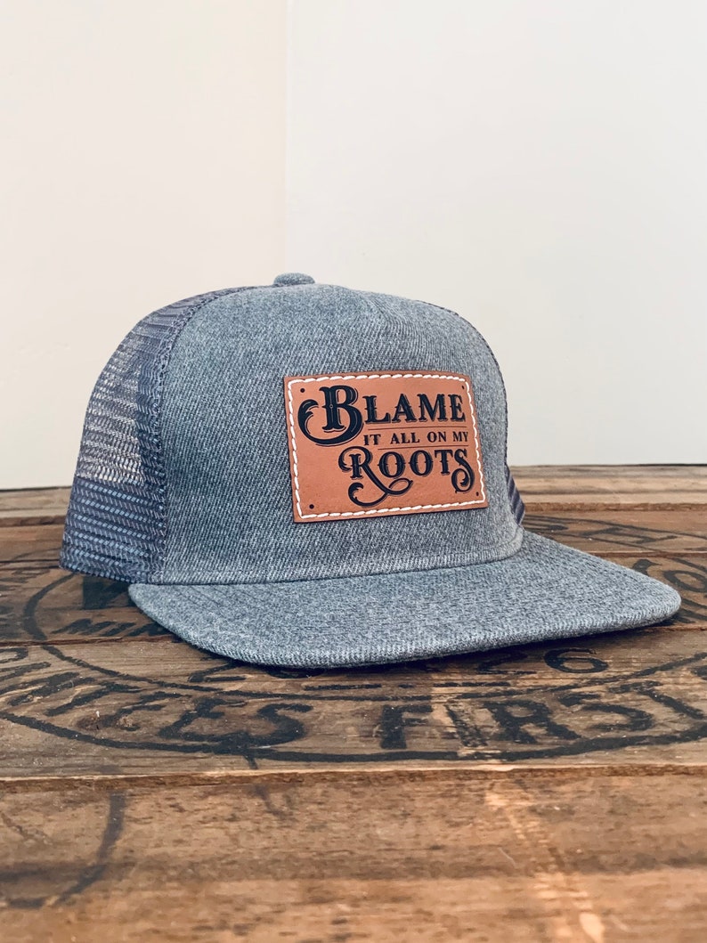 May include: A grey trucker hat with a mesh back and a flat brim. A rectangular tan leather patch is stitched to the front, featuring the black text "BLAME IT ALL ON MY ROOTS" in a decorative font with white stitching around the patch border.