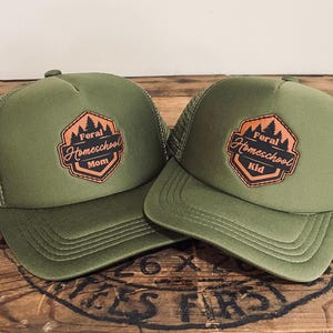 May include: Two olive green trucker hats. Each hat features a patch with the text "Feral Homeschool" and the words "Mom" and "Kid" respectively. The patches are in a brown and orange color scheme.
