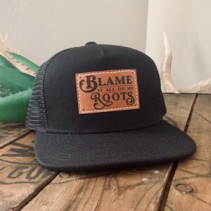 May include: A black trucker cap featuring a rectangular tan leather patch on the front panel. The patch is stitched with white thread and displays the black text "BLAME IT ALL ON MY ROOTS" in a decorative font style with a curved brim.