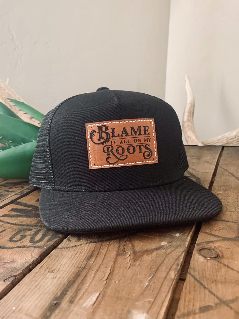 May include: A black trucker cap featuring a rectangular tan leather patch on the front panel. The patch is stitched with white thread and displays the black text "BLAME IT ALL ON MY ROOTS" in a decorative font style with a curved brim.