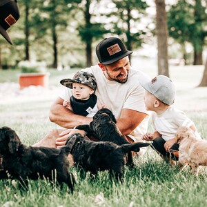 May include: A collection of baseball caps including a black mesh-back cap with a rectangular leather patch, a camouflage print cap, and a solid light grey cap. The black cap features the text "BLAME IT ON MY ROOTS" on the leather patch.