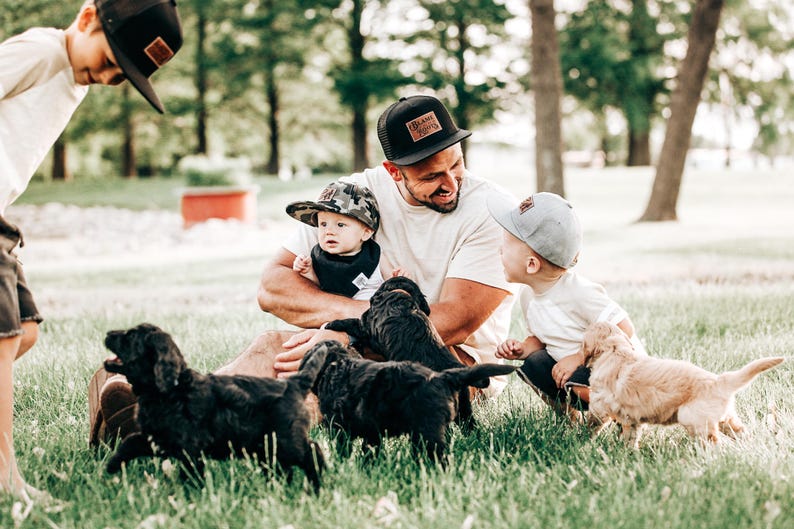 May include: A collection of baseball caps including a black mesh-back cap with a rectangular leather patch, a camouflage print cap, and a solid light grey cap. The black cap features the text "BLAME IT ON MY ROOTS" on the leather patch.