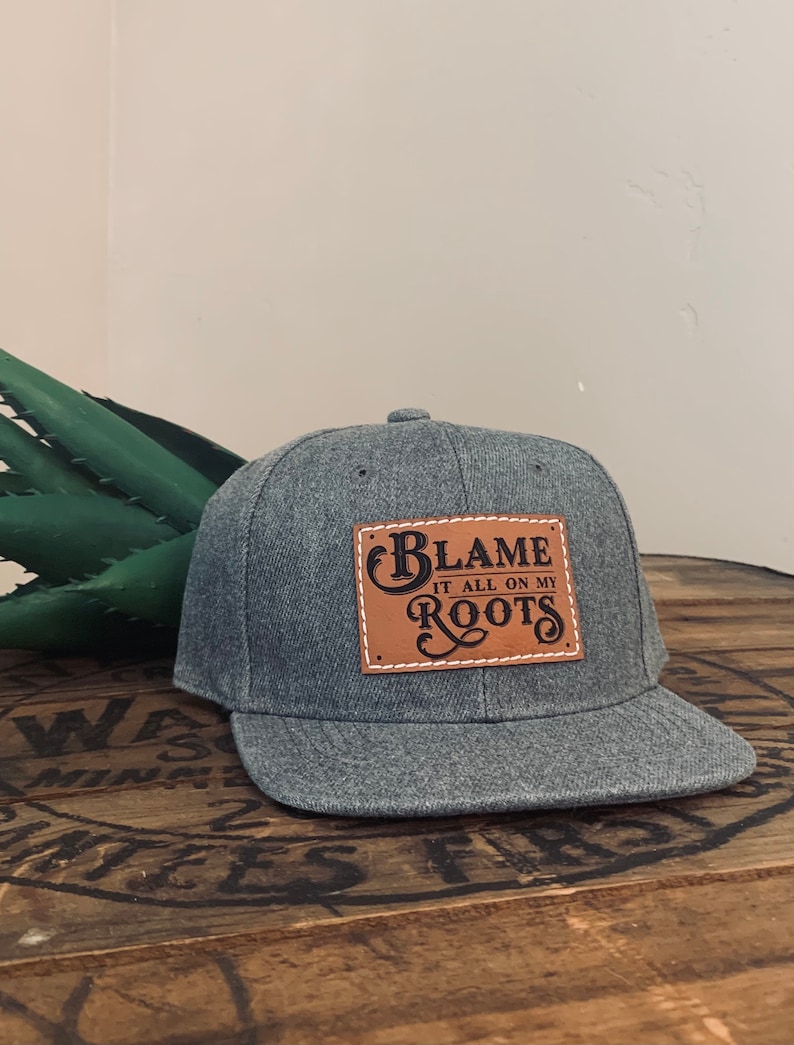 May include: A heather grey baseball cap featuring a rectangular tan leather patch stitched onto the front. The patch displays the black text "BLAME IT ALL ON MY ROOTS" in a decorative font, with white stitching around the patch border.