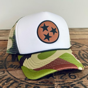 May include: A white trucker hat with a camouflage brim and a brown leather patch featuring three black stars inside a circle. The hat has a mesh back and a black rope detail.