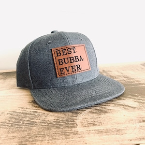 以下が含まれることがあります： 「BEST BUBBA EVER.」と書かれた茶色の革のパッチが付いた灰色のベースボールキャップ