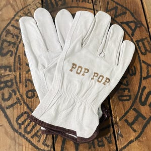 以下が含まれることがあります： 「POP POP」の文字が茶色で書かれた白い革製作業用手袋。手袋の袖口には茶色のトリムがあり、木製の表面に置かれています。