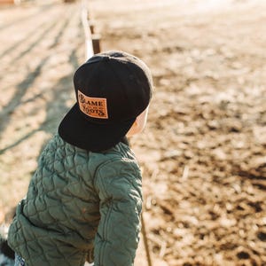 May include: A black baseball cap featuring a rectangular tan leather patch on the front. The patch is embossed with the text BLAME MY ROOTS in a stylised font. The cap is worn with an olive green quilted jacket that has a textured diamond pattern.