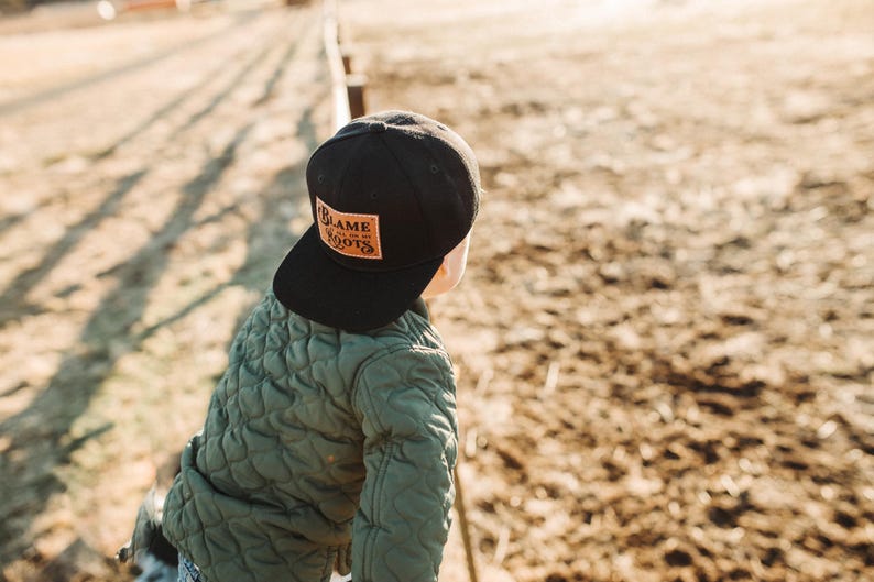 May include: A black baseball cap featuring a rectangular tan leather patch on the front. The patch is embossed with the text BLAME MY ROOTS in a stylised font. The cap is worn with an olive green quilted jacket that has a textured diamond pattern.