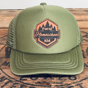 May include: Olive green trucker hat with a mesh back and a curved brim. The front features a patch with the text "Feral Homeschool Kid" in a black and orange design, with a forest graphic above the text.