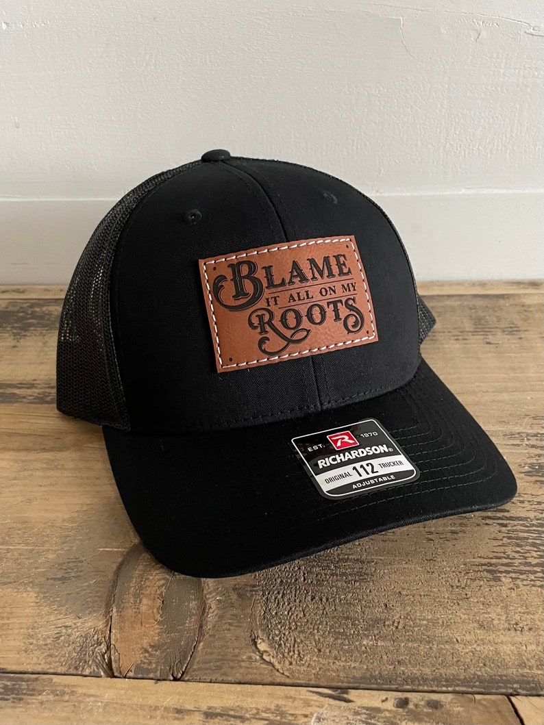 May include: Black trucker hat with a mesh back and a curved brim. The hat features a brown leather patch with the words "BLAME IT ALL ON MY ROOTS" in white lettering. The hat is adjustable and has a Richardson 112 tag.