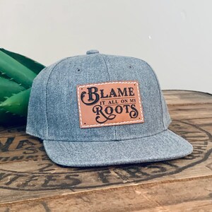 May include: A heather grey baseball cap with a flat brim and a rectangular tan leather patch stitched to the front. The patch features the black text "BLAME IT ALL ON MY ROOTS" in a decorative, stylised font with a prominent capital B and R.