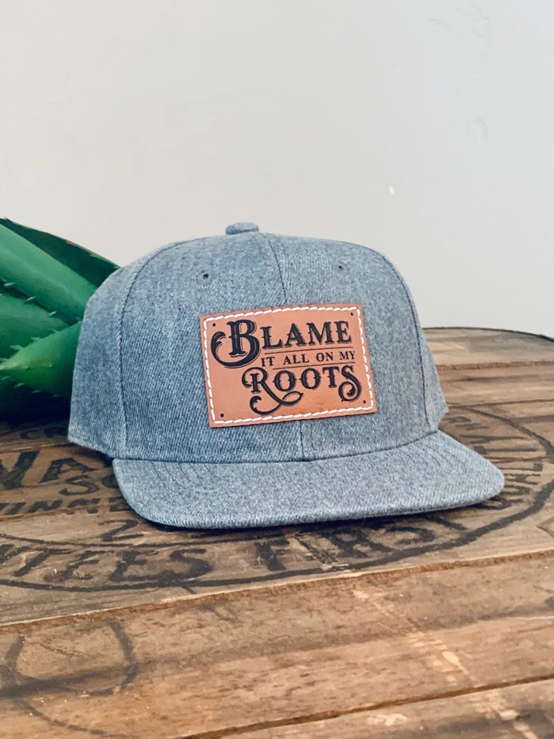 May include: A heather grey baseball cap with a flat brim and a rectangular tan leather patch stitched to the front. The patch features the black text "BLAME IT ALL ON MY ROOTS" in a decorative, stylised font with a prominent capital B and R.