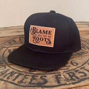 May include: A black baseball cap featuring a rectangular tan leather patch stitched onto the front. The patch displays the black text "BLAME IT ALL ON MY ROOTS" in a decorative font. The cap has a flat brim and a structured crown design.