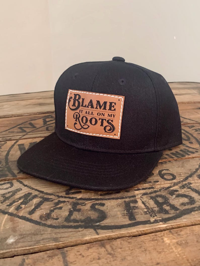 May include: A black baseball cap featuring a rectangular tan leather patch stitched onto the front. The patch displays the black text "BLAME IT ALL ON MY ROOTS" in a decorative font. The cap has a flat brim and a structured crown design.