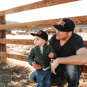 May include: Two black baseball caps with rectangular leather patches featuring the text "BLAME ROOTS". The caps have a structured crown and a flat brim. One cap is worn by an adult and the other by a child, both styled with casual clothing.