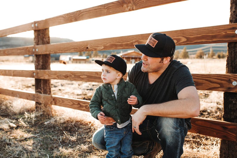 May include: Two black baseball caps with rectangular leather patches featuring the text "BLAME ROOTS". The caps have a structured crown and a flat brim. One cap is worn by an adult and the other by a child, both styled with casual clothing.