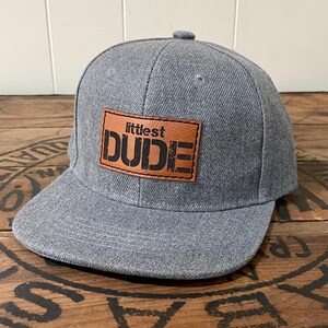 May include: Gray and brown baseball cap with the text "littlest DUDE" on the front.