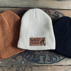 May include: Three knitted beanies in brown, beige, and black. The beige beanie has a brown leather patch with the text "WILD CHILD" and a cactus graphic.