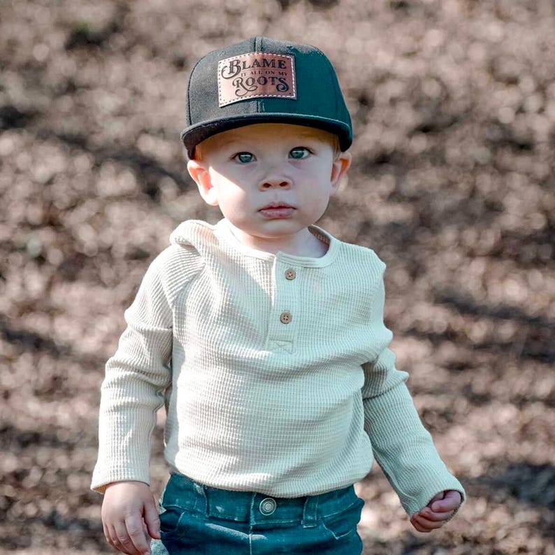 May include: A black baseball cap with a rectangular brown leather patch featuring the text "BLAME IT ALL ON MY ROOTS" in black lettering. The cap is paired with a long-sleeved, cream-coloured waffle-knit shirt with a three-button placket and dark jeans.