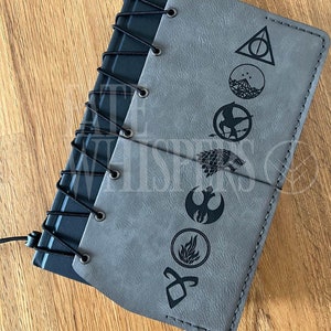 May include: A gray leather-bound journal with a black spiral binding. The cover features seven different symbols, including the Deathly Hallows symbol, the Mockingjay symbol, and the Star Wars Rebel Alliance symbol.