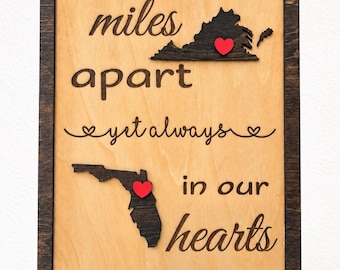 Könnte beinhalten: Ein rechteckiges Holzschild mit dunkelbraunem Rahmen und hellem Holzhintergrund. Es zeigt den Text miles apart yet always in our hearts sowie dunkel gebeizte Holzausschnitte von Virginia und Florida, die jeweils mit einem kleinen roten Herz versehen sind.