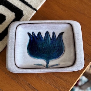 Op de afbeelding: Een klein, vierkant keramisch schaaltje met een verhoogde rand. Het schaaltje is wit met een blauw en groen bloemmotief in het midden.